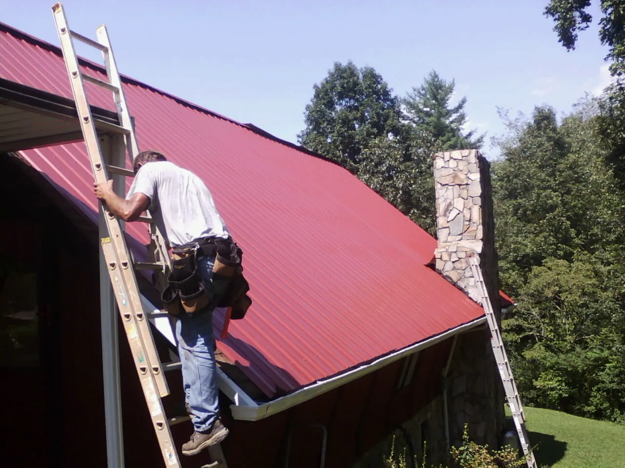 Roofer on a ladder beside a red metal roof and stone chimney — Soffit & Fascia Repair by Roofing Brothers in Red Bluff